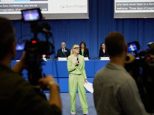 This image captures a conference scene. In the foreground, a camera operator is seen from behind, focusing on a woman speaking at the center. She is dressed in a vibrant green suit and holds a microphone, suggesting she is addressing the audience. In the background, a panel with four individuals is visible, two women and two men, with conference branding that reads "Zero Project" promoting a world with zero barriers. The setting is professional and conveys a sense of an event dedicated to inclusivity and perhaps issues related to accessibility and equality.
