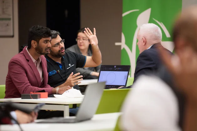 The photo shows a diverse group of individuals engaged in what appears to be a collaborative setting, likely an educational or professional environment. In the foreground, two men are conversing actively, with one man explaining something to the other and gesturing towards a laptop screen. Another person in the background seems to be raising her hand, perhaps to ask a question or contribute to the discussion. The presence of laptops suggests a modern, tech-oriented workspace or learning session. This setting exemplifies an inclusive atmosphere where people of different backgrounds are engaged in constructive dialogue and learning, advocating values of equality, tolerance, and mutual assistance.