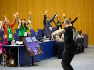 This image captures a scene inside a conference or meeting room. Several individuals are standing and raising their hands, possibly participating in a vote or expressing agreement. They exhibit a mix of apparent genders and potentially diverse backgrounds, symbolizing inclusion and collective decision-making. In the foreground, a person appears to be moving quickly, perhaps conducting the proceedings or facilitating the event. Their motion contrasts with the stationary posture of others. A large screen, mimicking the room's activities, indicates the use of technology for visibility or recording purposes. The setting evokes a spirit of democracy, collaboration, and active engagement, embodying themes of equality and justice.